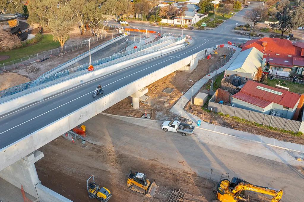 A motorcycle rides over a bridge which spans a construction area, in a urban-rural setting.