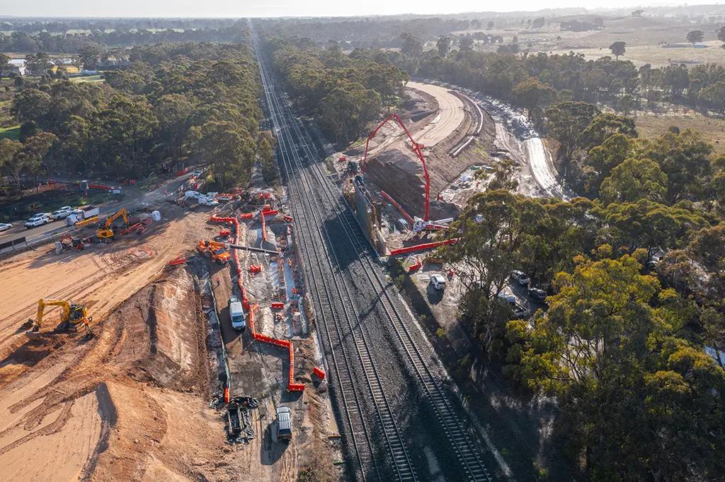 A construction site in a rural area. A rail line runs through the centre of it. A road is being constructed either side of the railway line. Machinery and vehicles are dotted across the site. There are mountains on the horizon.