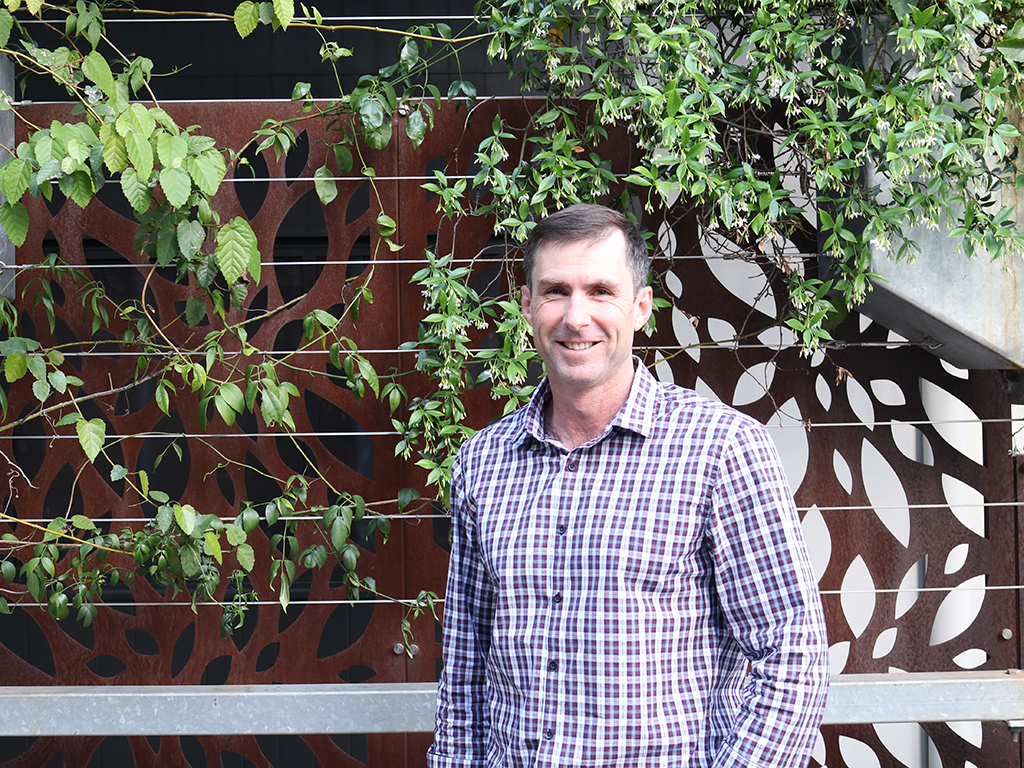 A man standing facing the camera, smiling, in front of a green vine