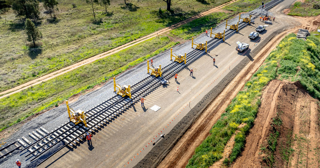 A series of bright yellow spider-like machines straddle a shiny new section of rail, in a rural setting.