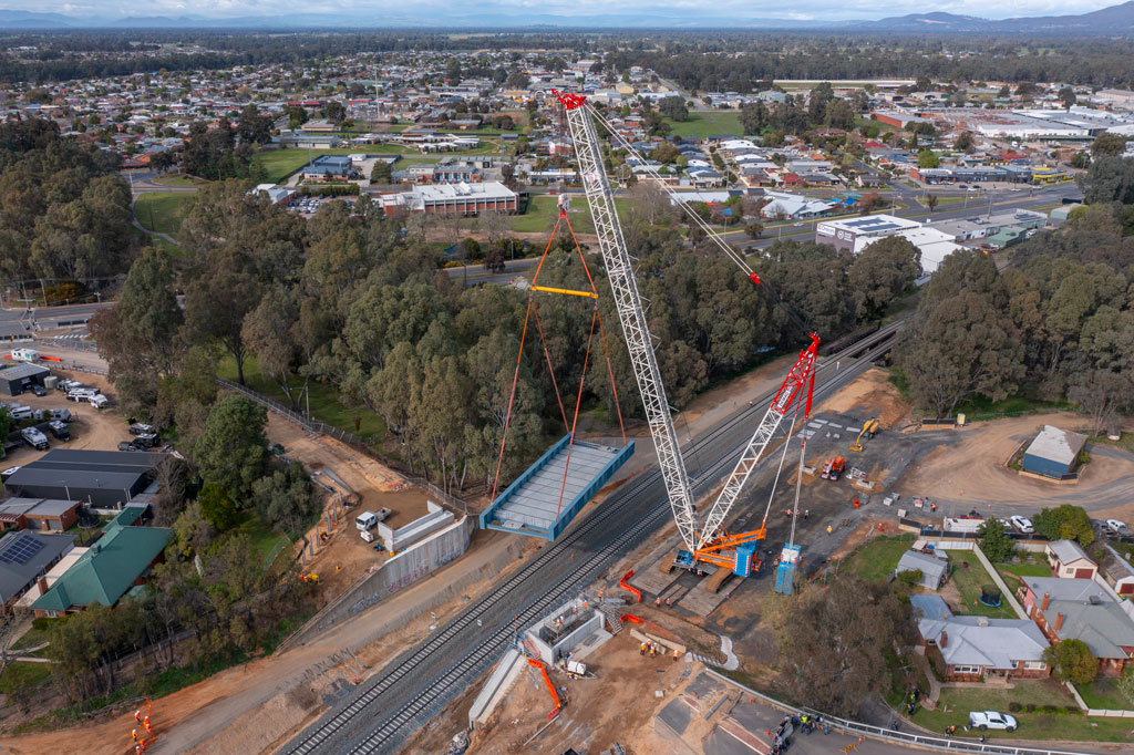 A huge crane lifts a massive bridge span into place over two tracks of rail. A township and mountains can be seen in the background, which shows the scale of the crane, as it towers over the landscape.