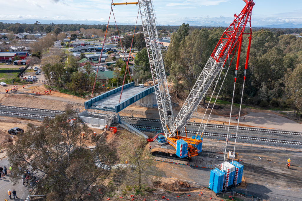 A view, taken high in the air from a drone, shows a very tall crane towering over the landscape. It is lifting a massive blue bridge span into place over two railway tracks. Here, it lowers the bridge into place, ready for it to be secured to the bridge supports.