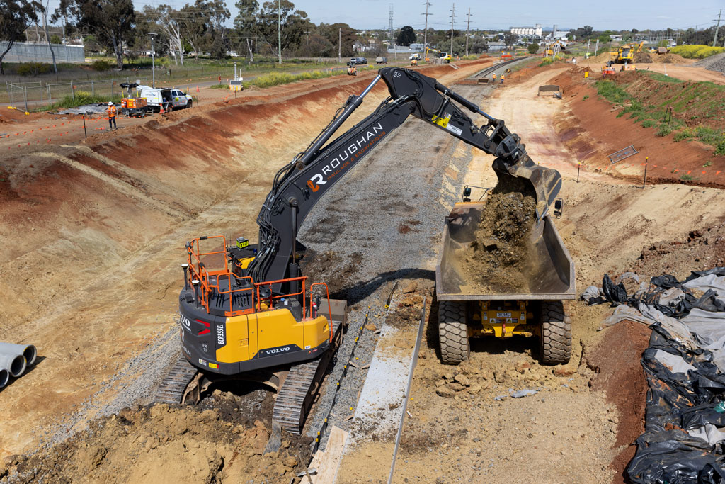 A construction site. An excavator's long arm deposits dirt into a tip truck. Noth machines are next to a rail line which is covered in ballast.