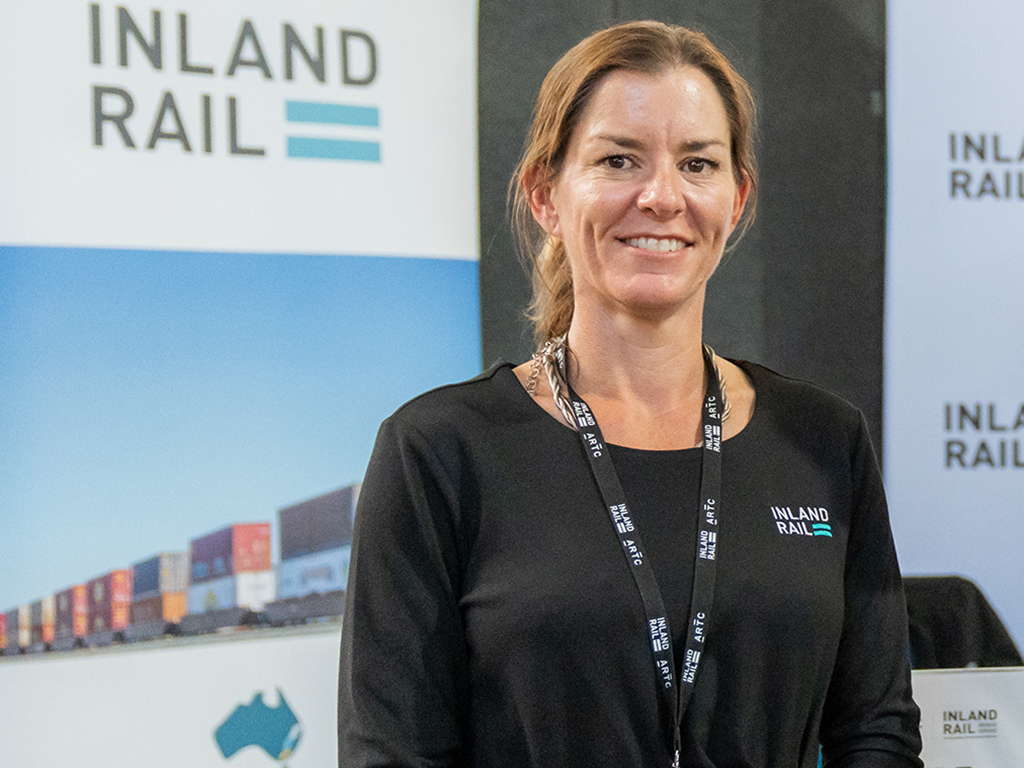 A woman standing facing the camera, smiling, with a banner displaying the Inland Rail logo