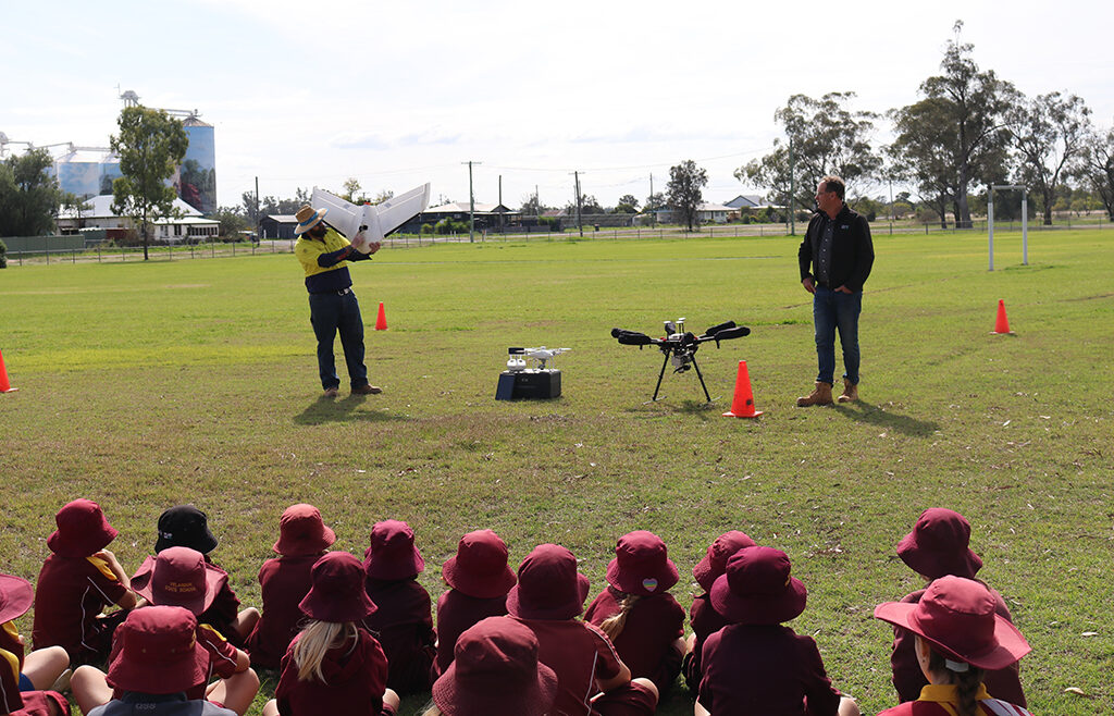 Two men standing in a field with drone equipment in front of a seated group of students