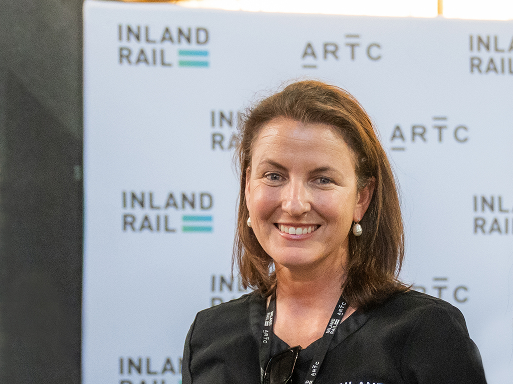 A woman standing facing the camera, smiling, with an Inland Rail banner in the background