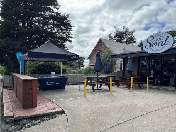 Outside a cafe, a seating area holds a tend with a desk under it. There are signs on the tent and desk saying ARTC Inland Rail, and information flyers on the table.