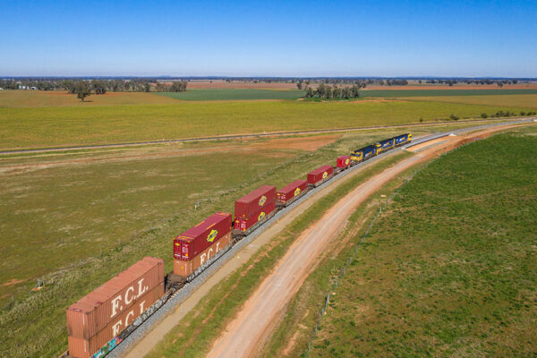 A train travels through a rural setting. The train's carriages hold two containers staked on each other.