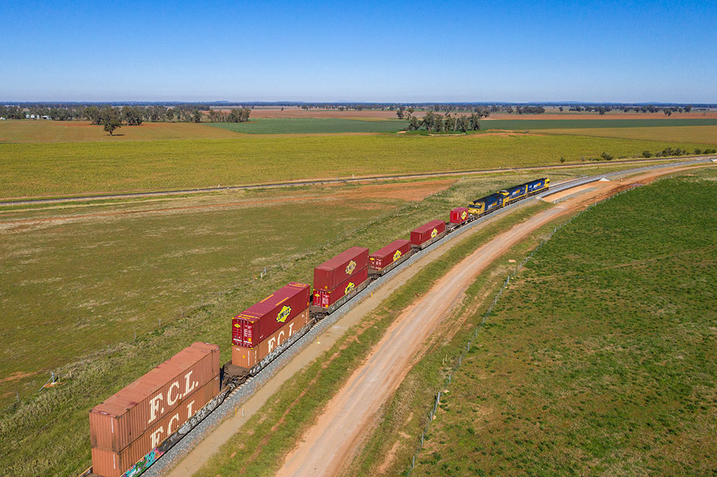 A train travels through a rural setting. The train's carriages hold two containers staked on each other.