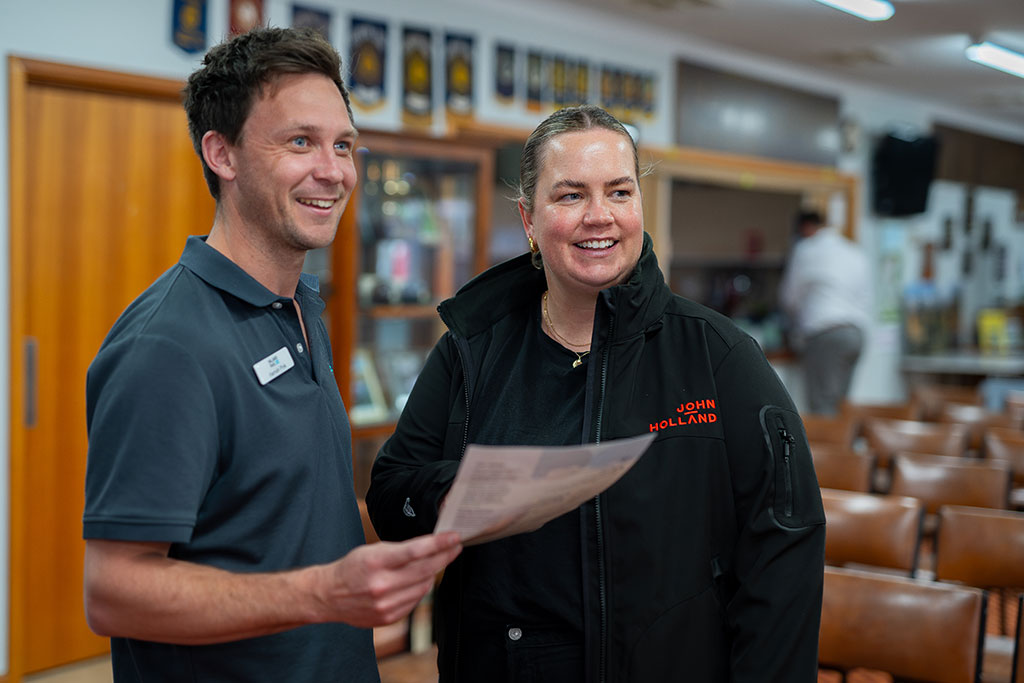 Two people stand in a room with chairs set up in rows behind them. One person holds a sheet of paper, and is wearing an Inland Rail name badge. The other person is wearing a jumper with John Holland brand name on it.