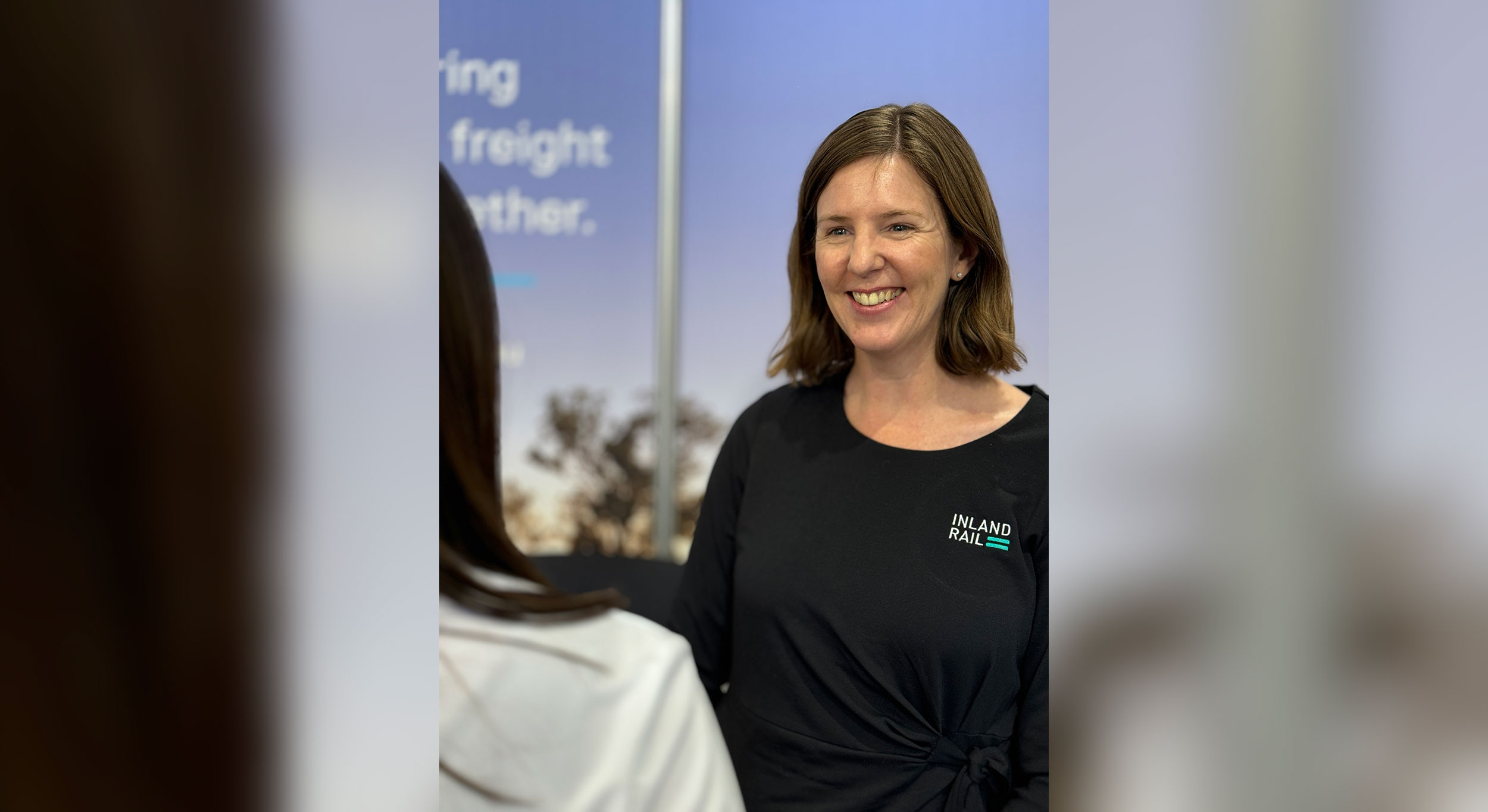 A woman standing facing the camera, smiling, wearing a shirt with the Inland Rail branding