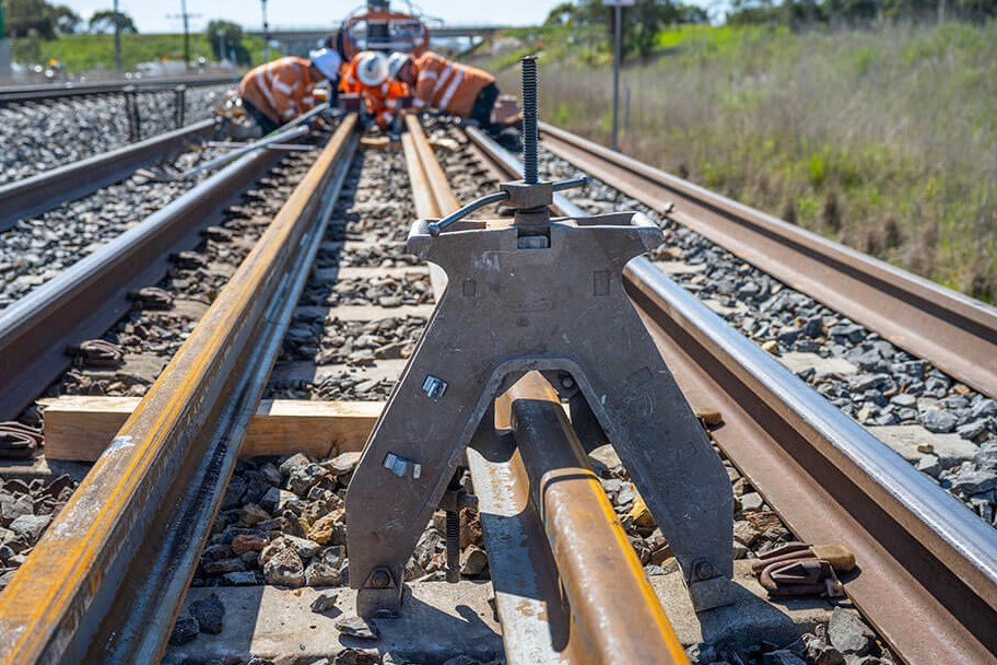 A railway track under construction. Workers can be seen in the background, crouching on the line. A metal pieces of equipment is in the foreground.