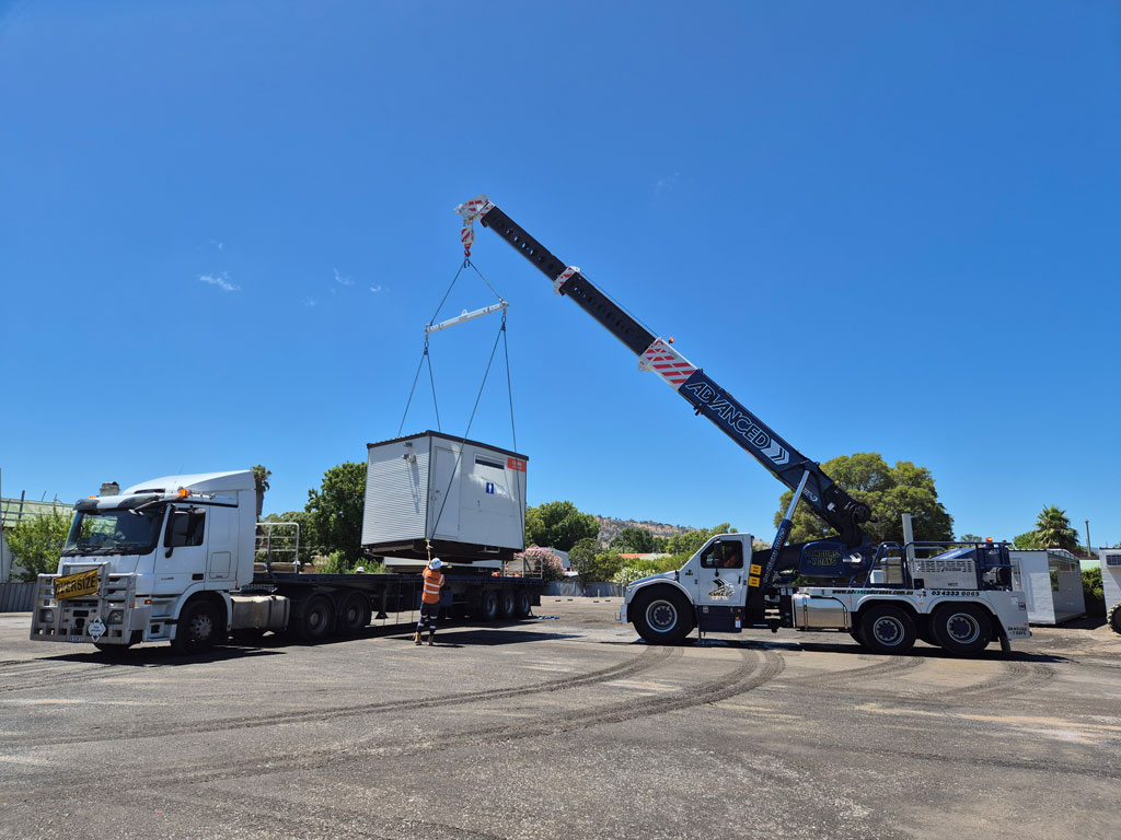 A crane lifts a building from the back of a truck in a large paved yard.