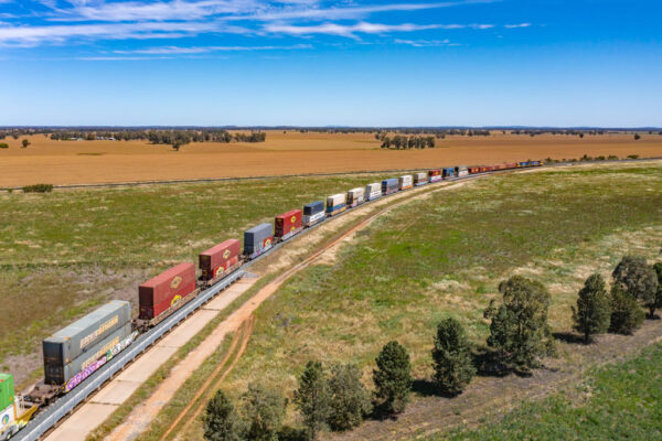 A train travels into the distance in a rural setting. The each train trailer carries two shipping containers, stacked on top of each other.