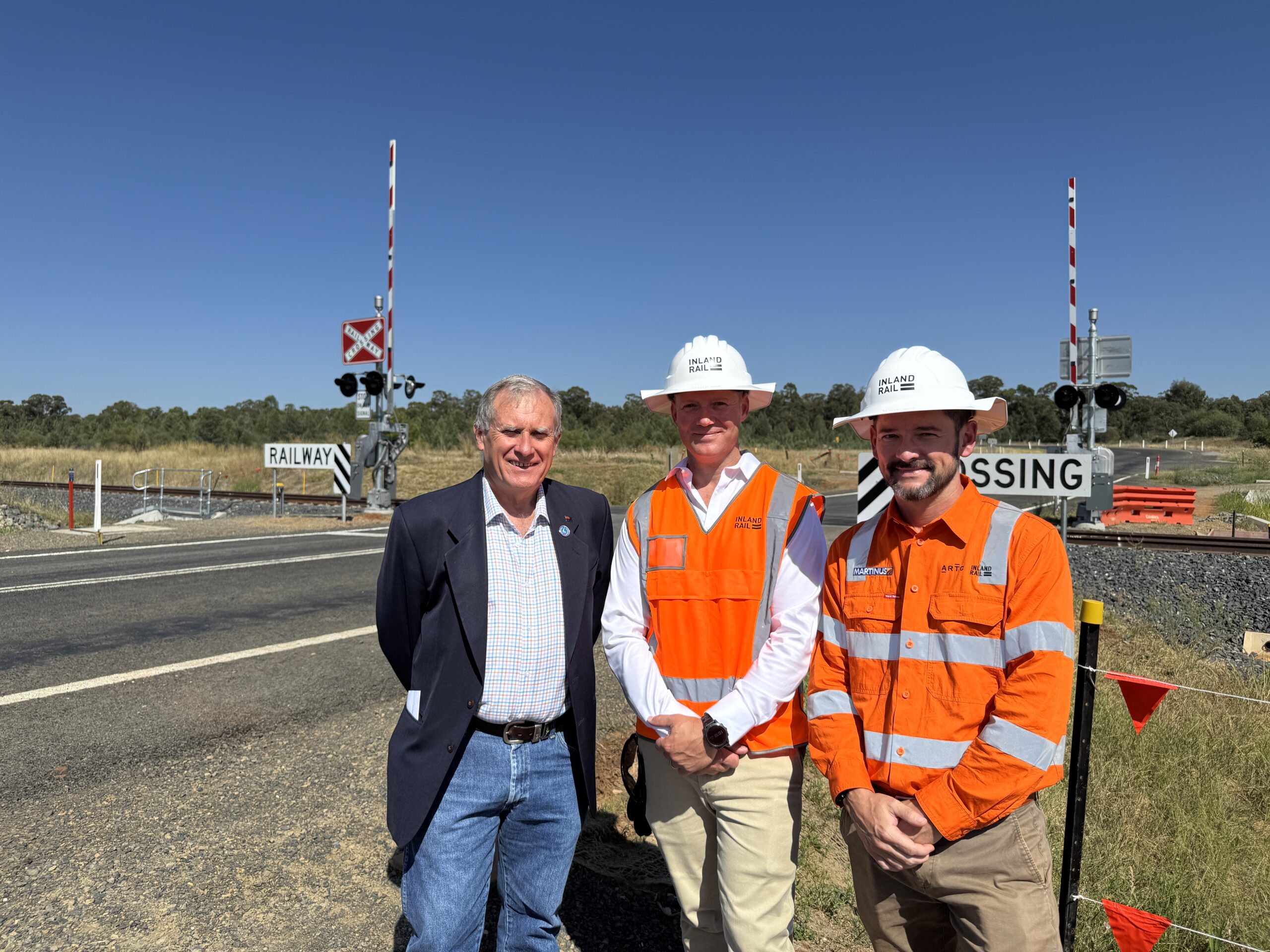 Three men, two in high visibility vests, look at a train level crossing, where a road is intersected by a rail line. Boom gates, flashing lights and stop signs are at the intersection.