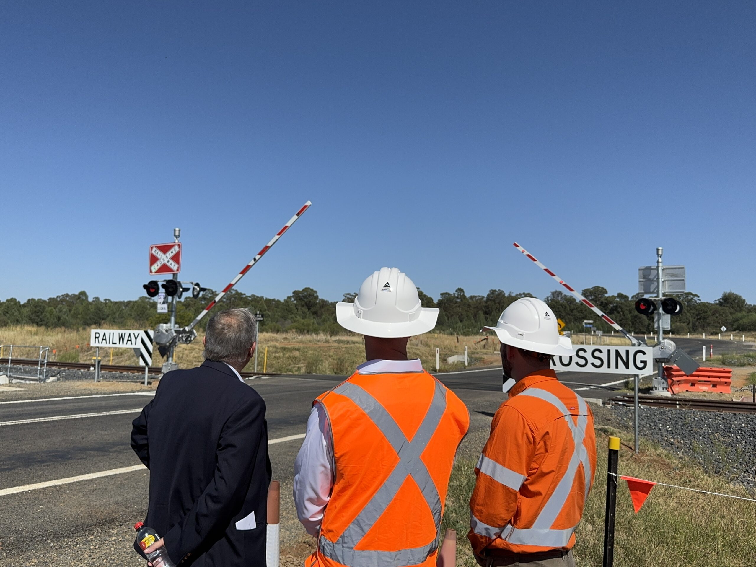 Three men, two in high visibility vests, look at a train level crossing, where a road is intersected by a rail line. Boom gates, flashing lights and stop signs are at the intersection.