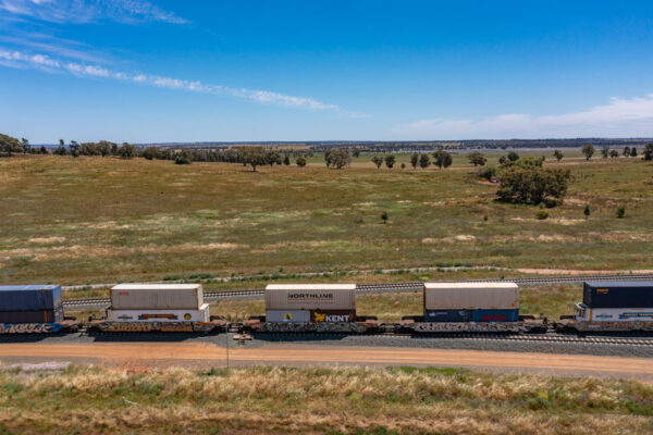 A train travels into the distance in a rural setting. The each train trailer carries two shipping containers, stacked on top of each other.