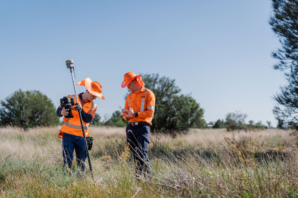 Two workers in high visibility workwear standin a rural field, holding survey equipment.