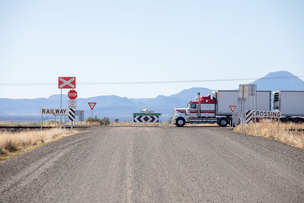 A road leads to a rail track. Signs saying Railway and Stop are positioned in front of the track, next to the road, in a rural setting.