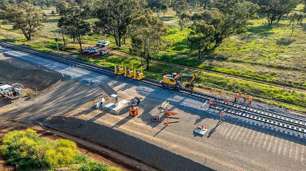 Some bright yellow spider-like machines straddle a shiny new section of rail, in a rural setting, with a yellow machine, construction crew and material around the track