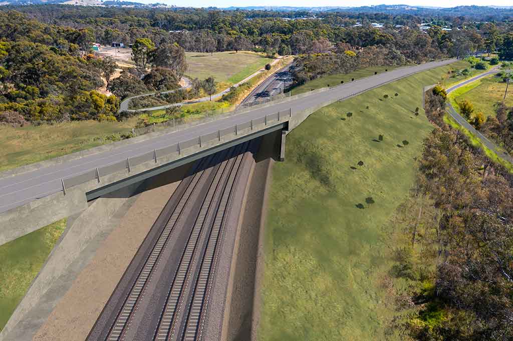A road bridge crosses over a railway corridor in a rural setting.