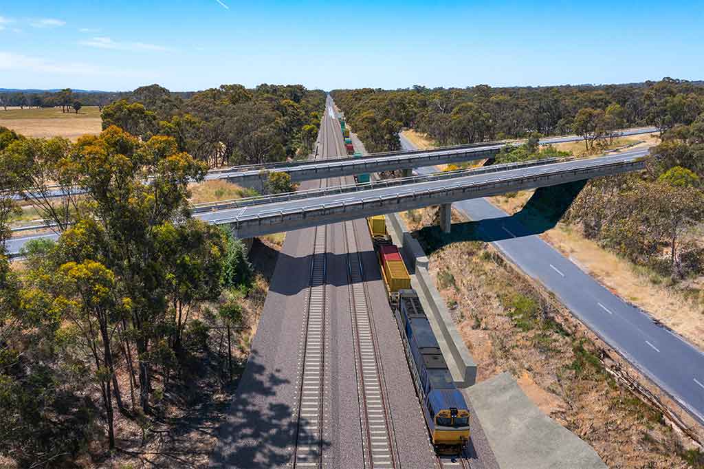 A three-track railway corrdor runs alongside a rural road in a bushland setting. Two road bridges cross over the railway and the road.