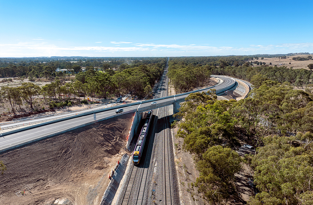 A curved road leads to a bridge over a railway corridor, in a rural landscape.
