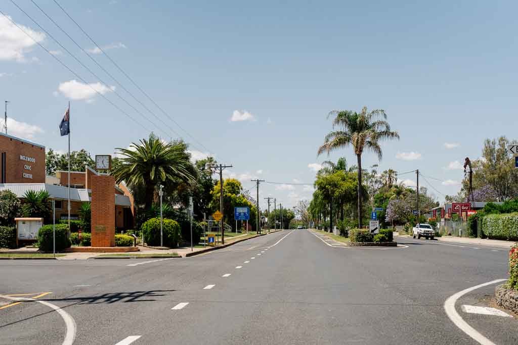 A town centre with a main street, with palm trees down the centre strip. A brick civic building is to one side of the road.