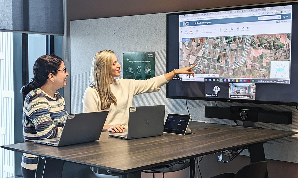 Two women with laptops standing at a desk with one woman pointing at a large screen displaying an aerial map