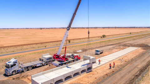 A crane lines up a row of concrete culverts, small bridge-like concrete structures, in a line in a rural setting.