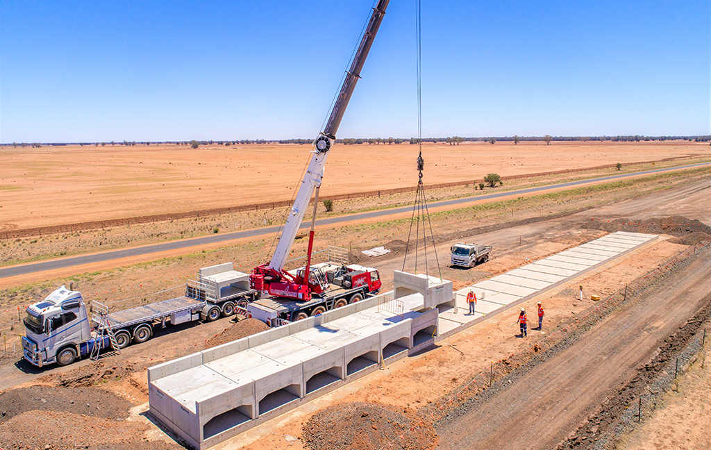 A crane lines up a row of concrete culverts, small bridge-like concrete structures, in a line in a rural setting. 