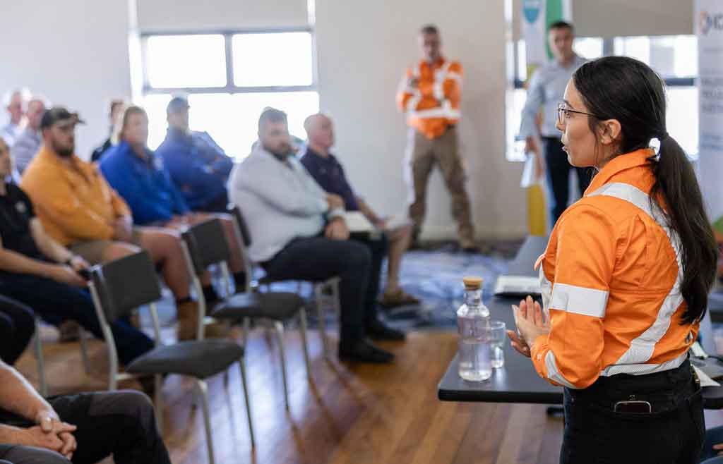 A person dressed in high visibility workwear speaks to a room full of people.