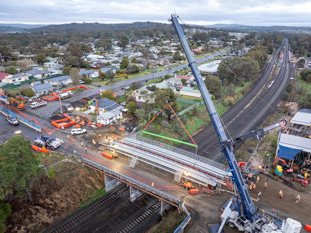 A crane lifting a bridge section over railway tracks in a residential area, with workers and construction equipment on site.