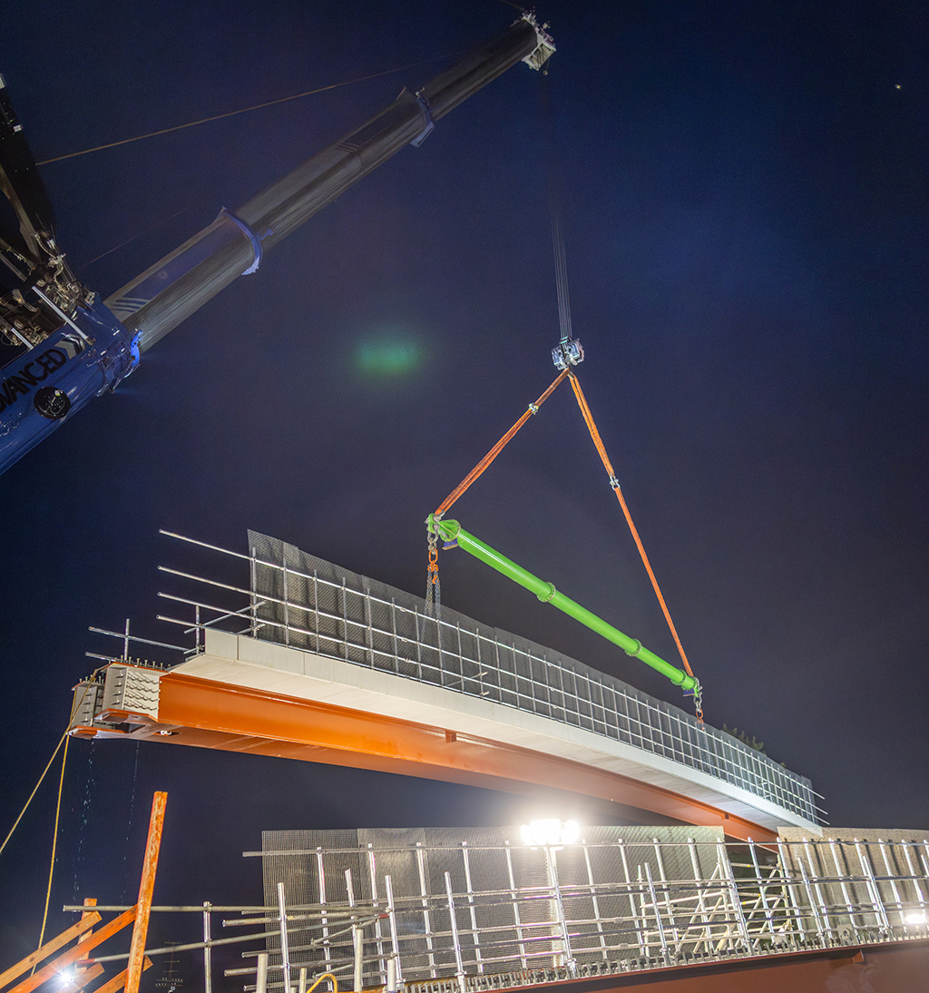 A crane lifting a bridge section at night, with construction materials and scaffolding illuminated by bright lights.