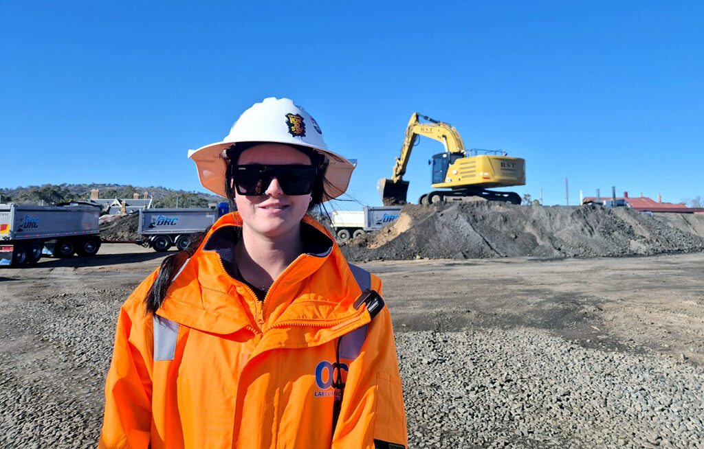 A construction worker in safety gear standing near an excavator and trucks at a construction site.