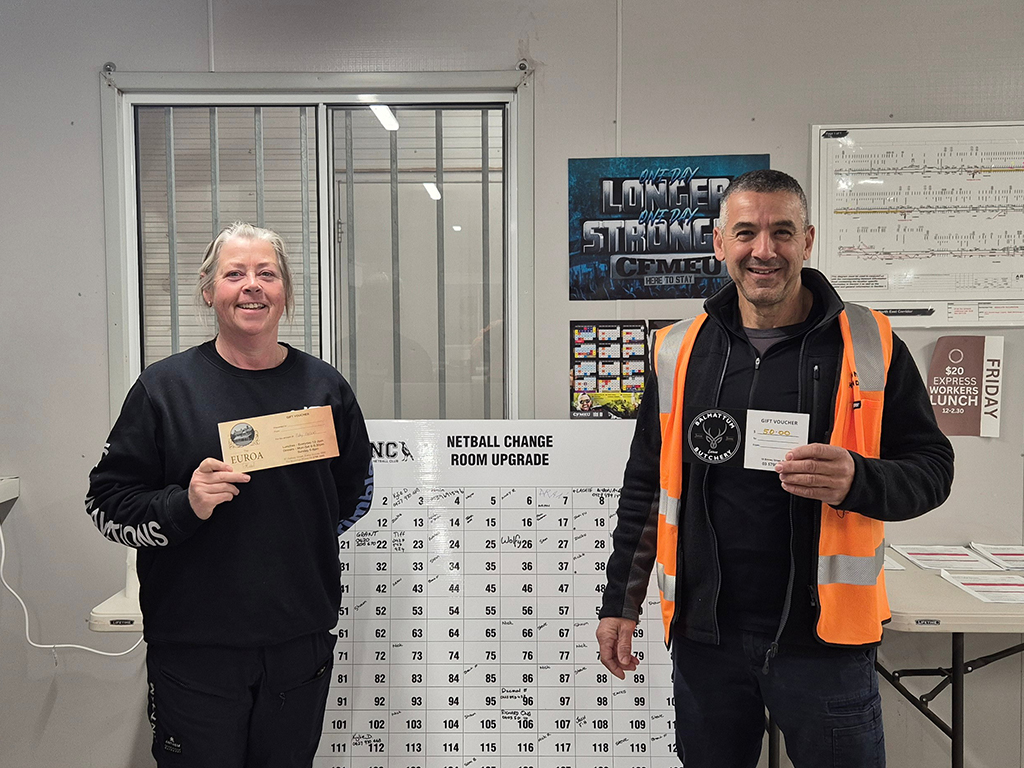 Two people holding gift vouchers in front of a 'Netball Change Room Upgrade' board with numbered grid and posters on the wall behind them.