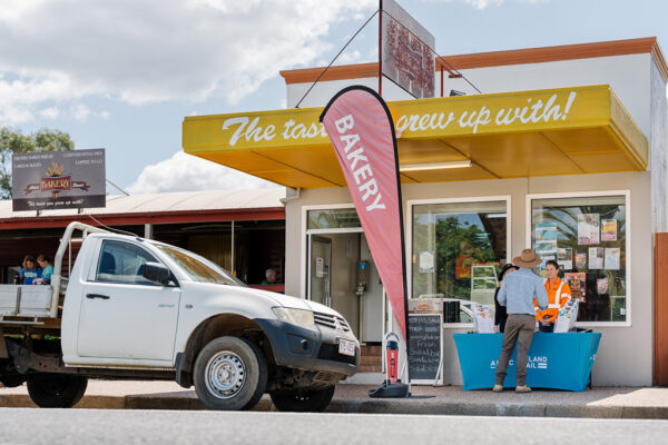 A bakery with a yellow awning and red 'BAKERY' sign, a white pickup truck parked in front, and two people near a table outside.