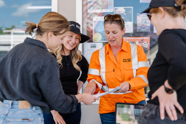 Four people standing together, one in an 'INLAND RAIL' high-visibility jacket holding documents, with a bulletin board in the background.