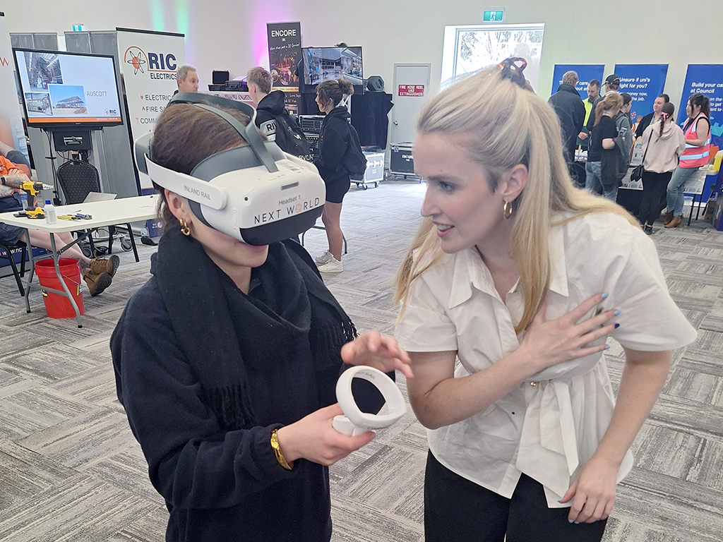 At a technology exhibition, a person uses a 'NEXT WORLD' virtual reality headset while another person provides guidance. Booths and attendees are visible in the background.