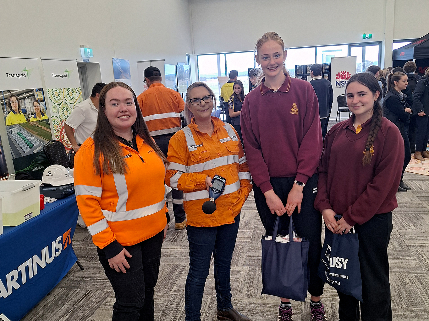 Group of four individuals at an industry expo, wearing high-vis and maroon uniforms, with booths in the background.