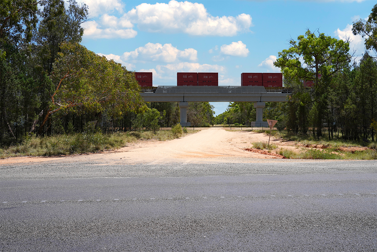 A concrete structure with a train running on it stands on concrete pylons on either side of a dirt road. The dirt road leads to a bitumen road, in a bushland setting.