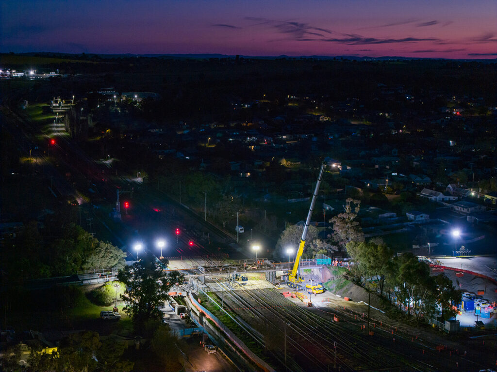Aerial view of a nighttime construction site with a crane and railway tracks, illuminated by bright lights.