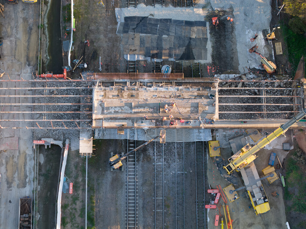 Aerial view of a bridge being demolished over railway tracks, with cranes, scaffolding, and workers visible.