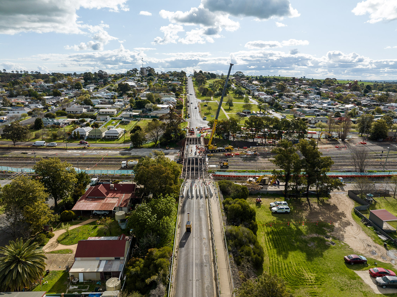 Aerial view of a bridge being demolished over railway tracks, with heavy machinery including a crane and construction vehicles present. The road leading to the bridge is blocked off, and the site is surrounded by residential houses, trees, and green spaces.
