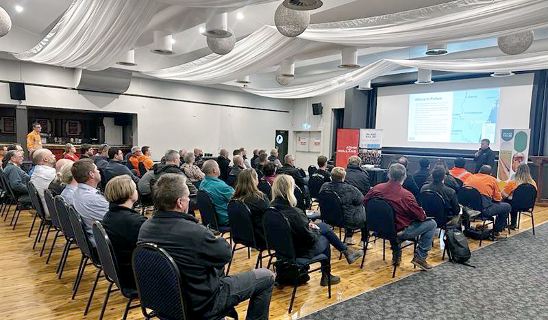 Group of people sitting in a room listening to a presentation with a man speaking with a microphone at the front with a slide being presented on a screen.