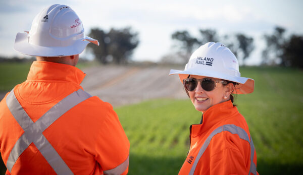 Two construction workers in orange high-visibility jackets and white hard hats standing in a grassy field, with trees in the background.