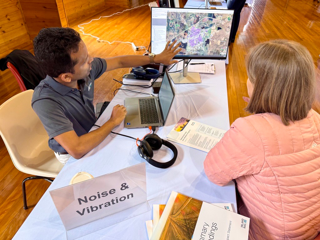 Two people sit at a desk. One is pointing to a computer screen that has a map on it while the other person looks on.