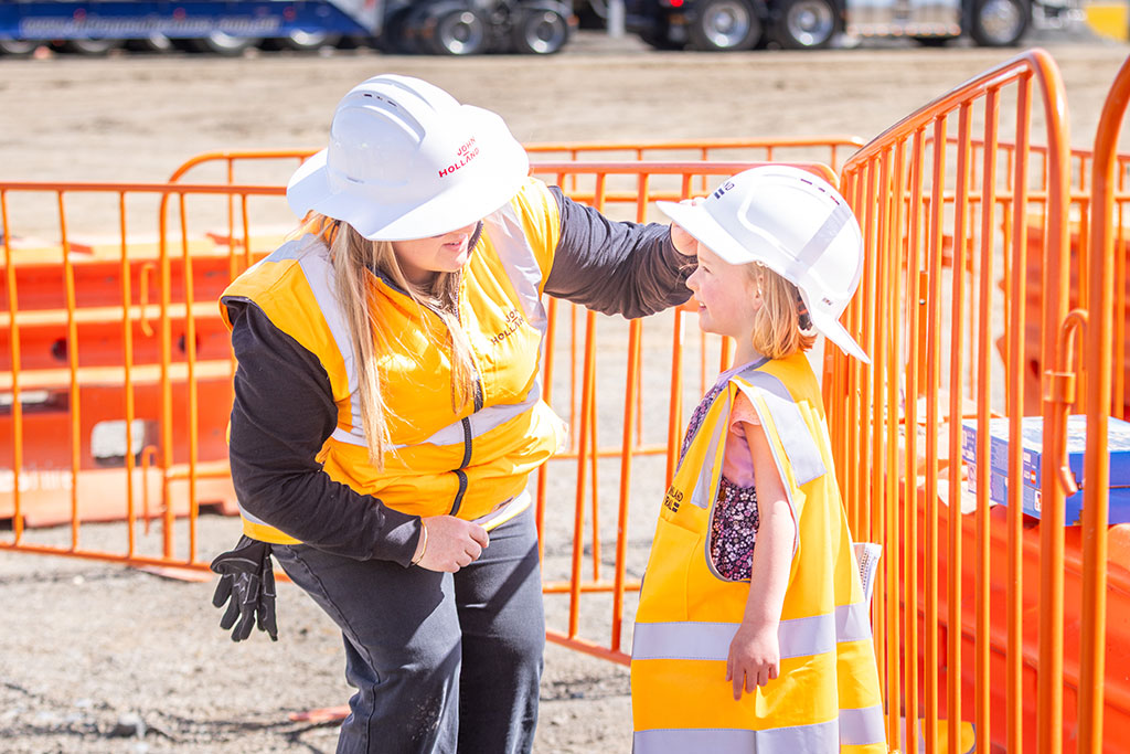A person in high visibility safety gear at a worksite bends down to adjust the hard hat of a young girl wearing a high visibility vest. The young girl is smiling.