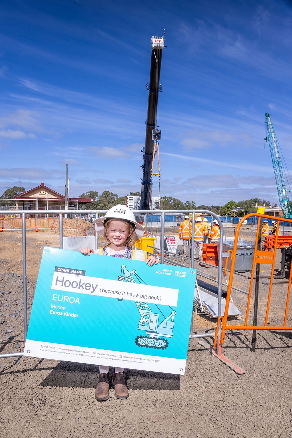 A child wears a hard hat and holds a sign up with the name of a crane, with a worksite and a crane behind her.