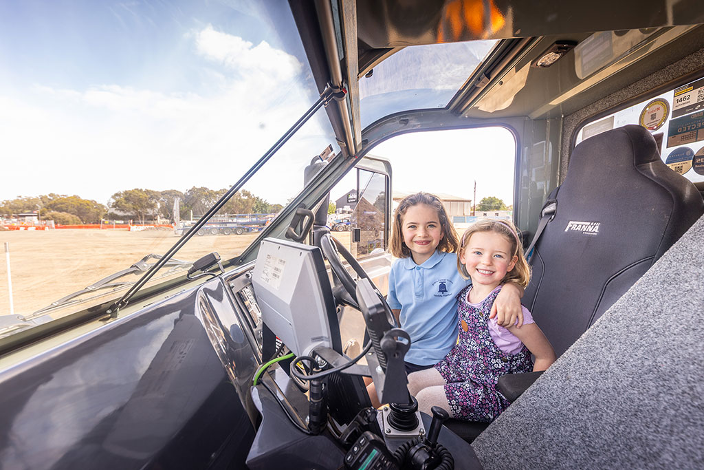 Two young children sit in the cab of a truck on a worksite.